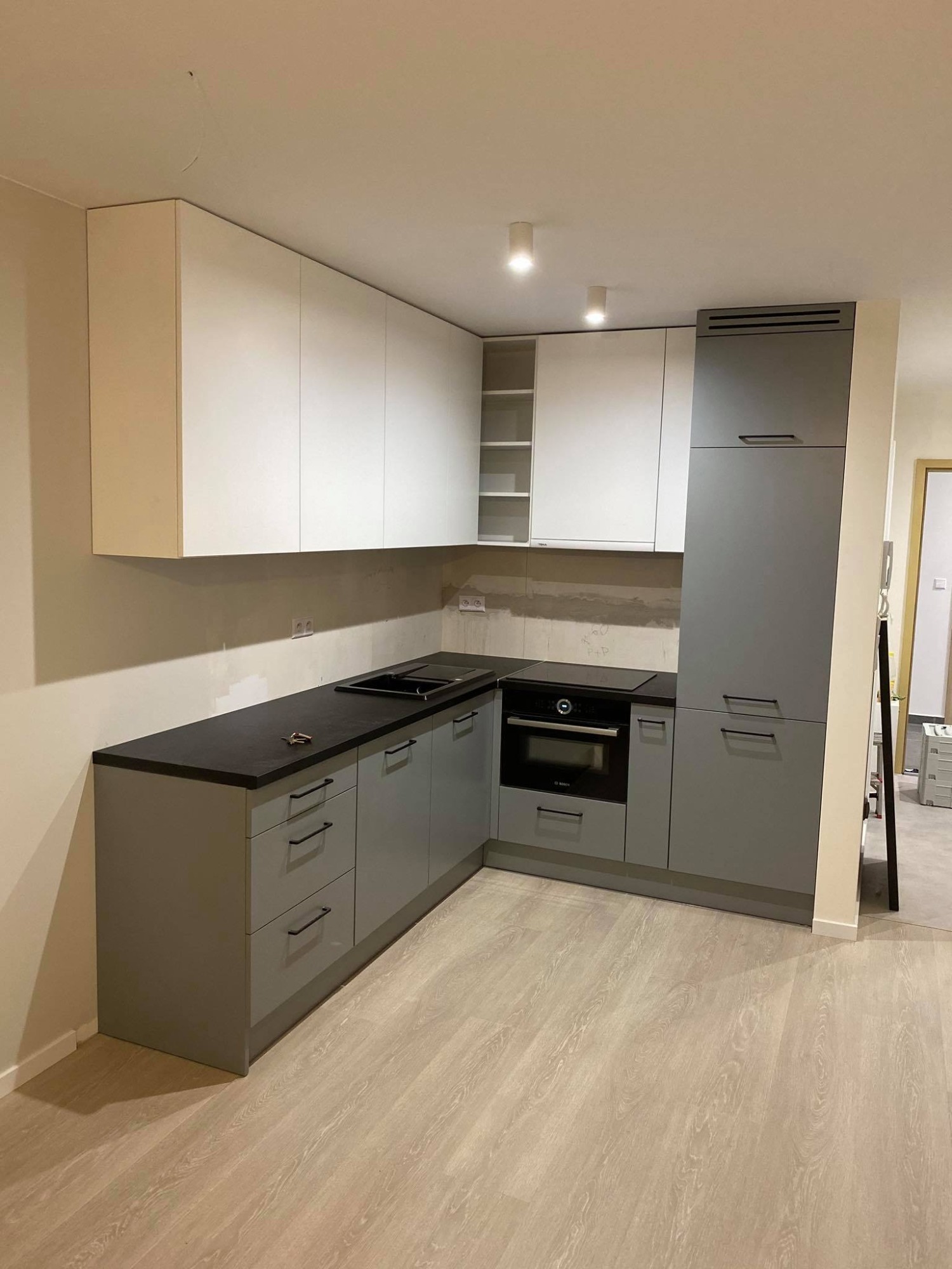 Nowoczesna kuchnia w odcieniach bieli i szarości z czarnym blatem, jeszcze w trakcie montażu, widoczne niedokończone ściany nad blatem.