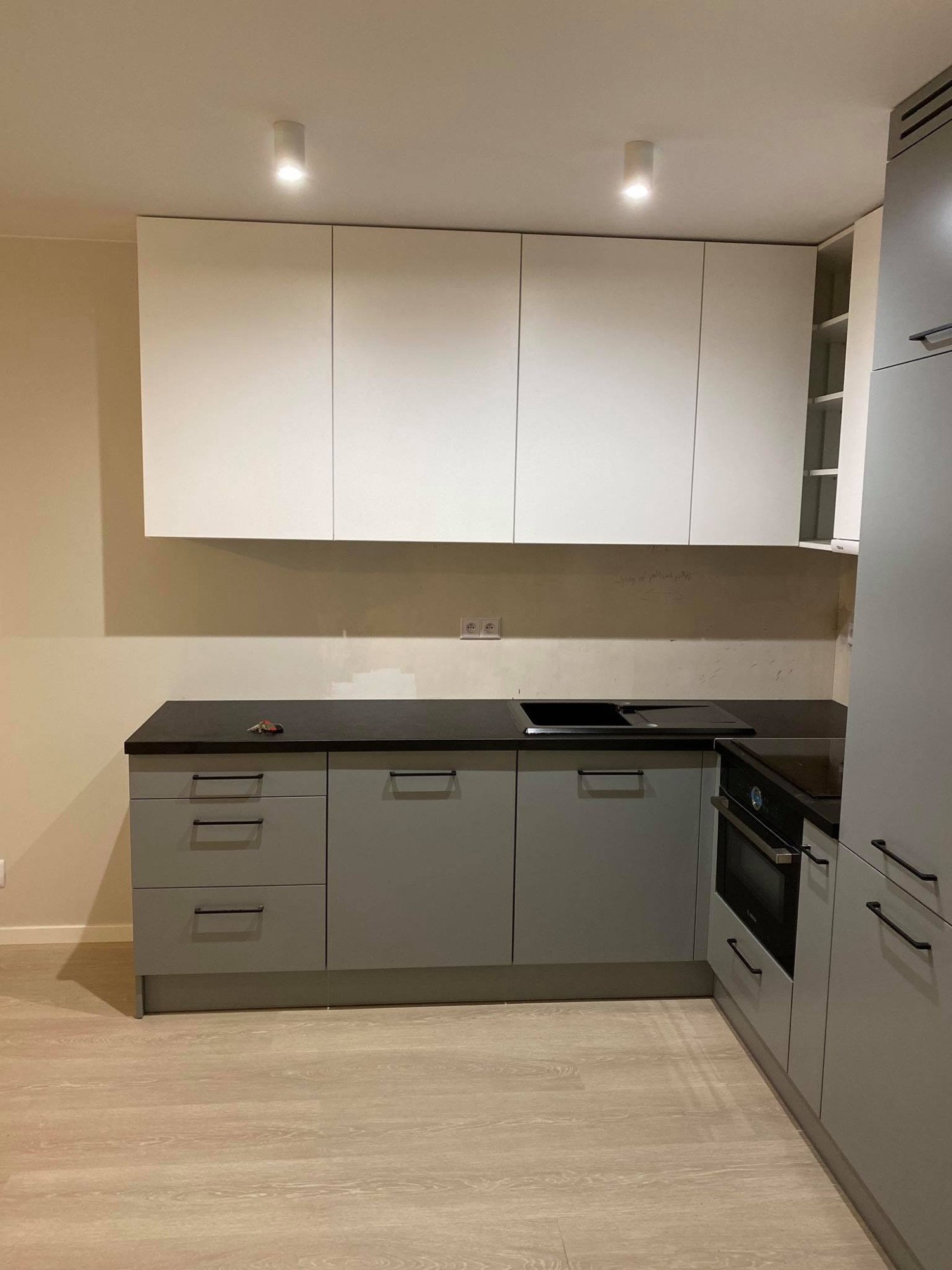 Szara zabudowa kuchenna z białymi szafkami wiszącymi, czarnym blatem i wbudowanym piekarnikiem; klucze leżą na blacie, widoczne gniazdka elektryczne na ścianie.