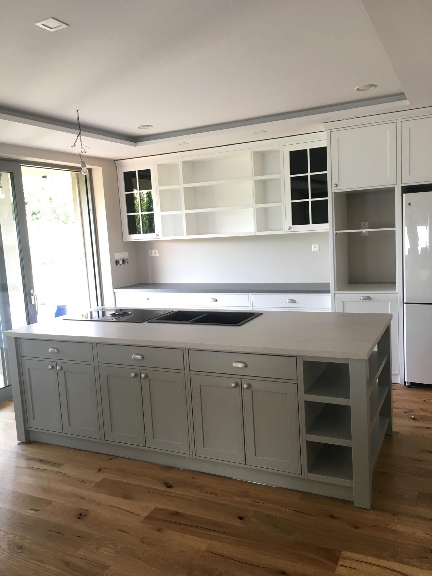 Jasna kuchnia z wyspą w Rudniku, szare fronty szafek dolnych i białe górnych, drewniana podłoga i duże okno balkonowe.