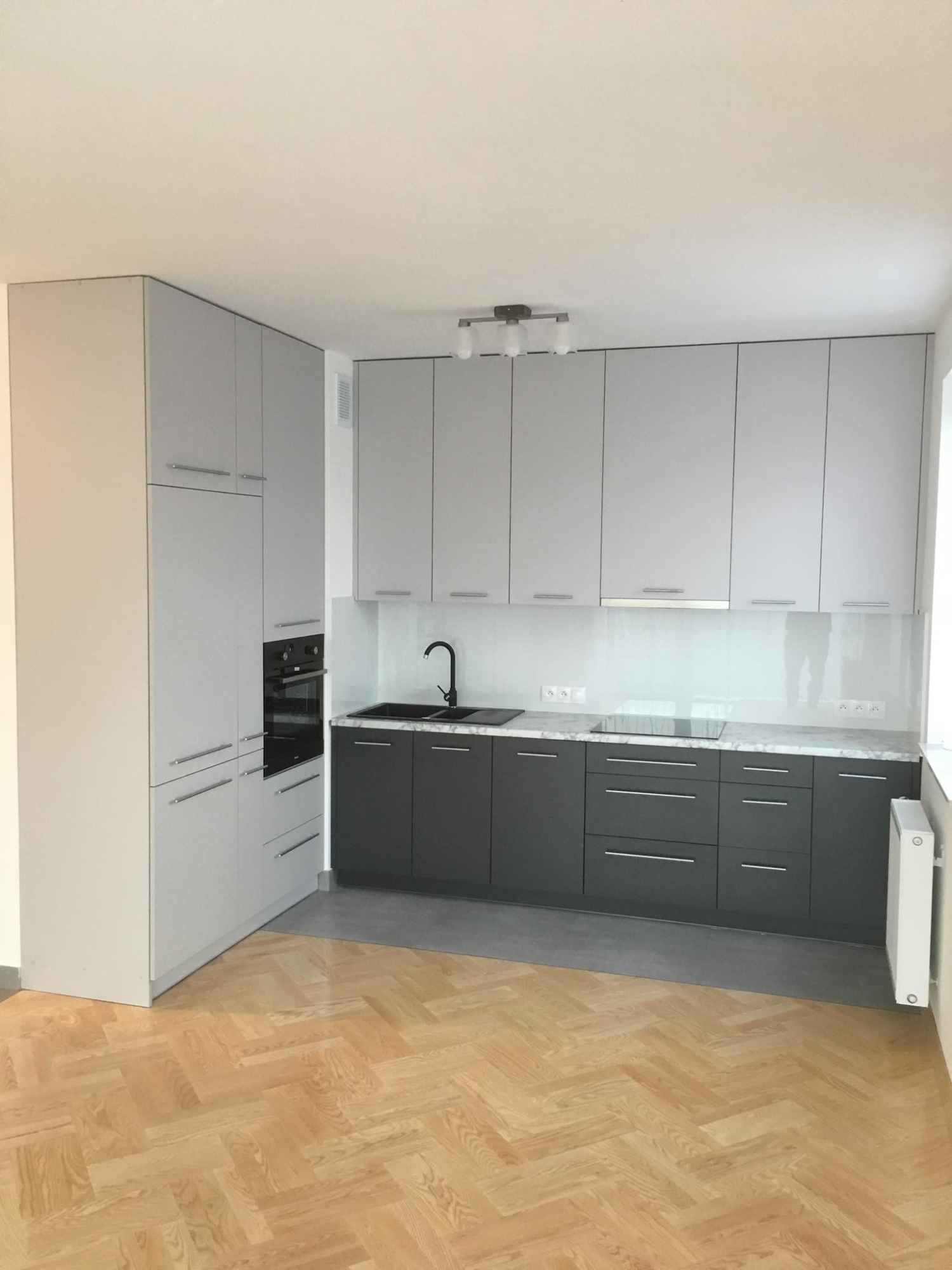Nowoczesna kuchnia z szarymi szafkami górnymi i grafitowymi dolnymi, blatem imitującym marmur, czarnym zlewem i oświetleniem sufitowym, na podłodze parkiet w jodełkę.