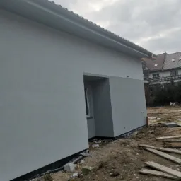 Świeżo otynkowana, szara elewacja domu jednorodzinnego w trakcie wykańczania, widoczny fragment okna, orynnowanie i teren wokół budynku z resztkami materiałów budowlanych.