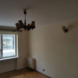 Pusty pokój przed remontem: parkiet, okno z widokiem na ulicę, grzejnik, żyrandol w stylu retro i kinkiet na ścianie.