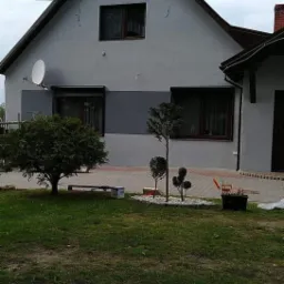 Dom jednorodzinny z szarą elewacją, widoczny fragment ocieplenia w postaci ciemniejszego pasa, antena satelitarna na elewacji, okna z brązowymi ramami i zadbany ogród z niskimi drzewkami.