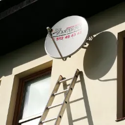 Biała antena satelitarna z logo firmy 'Słupski Serwis Antenowy' zamontowana na elewacji budynku, obok okna, z widoczną drabiną opartą o ścianę.