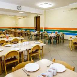 Sala jadalna z ustawionymi stołami dla dzieci, nakryta zastawa stołowa i kubki, ściany ozdobione poziomymi pasami w kolorach pomarańczowym, zielonym i niebieskim.