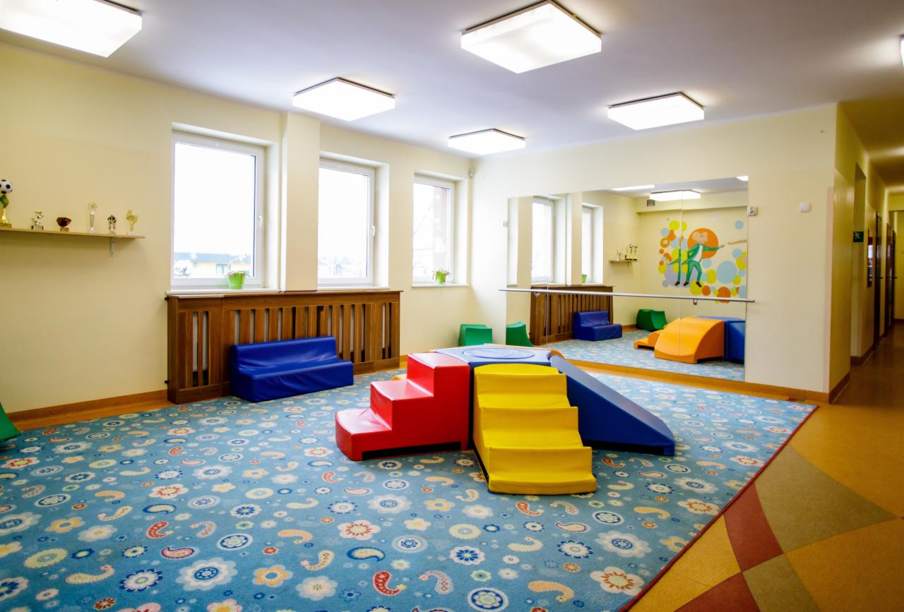 Jasne, przestronne wnętrze sali zabaw z kolorowymi meblami modułowymi dla dzieci, wzorzysty dywan, lustro na ścianie i półka z trofeami.