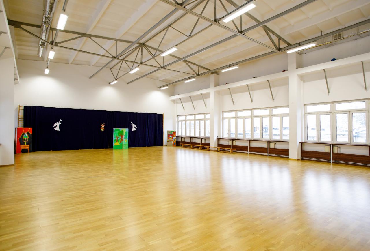 Duża, jasna sala z drewnianą podłogą, scena z ciemnoniebieską zasłoną ozdobioną białymi aniołami i kolorowymi dekoracjami, rząd okien z drewnianymi parapetami, białe ściany i sufit z widoczną...