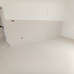Wróbel Dywan-Monter - Jasne, puste pomieszczenie z beżową, bezspoinową posadzką żywiczną o nakrapianej fakturze, przechodzącą płynnie w cokoły ścienne. Widoczne gniazdka elektryczne.