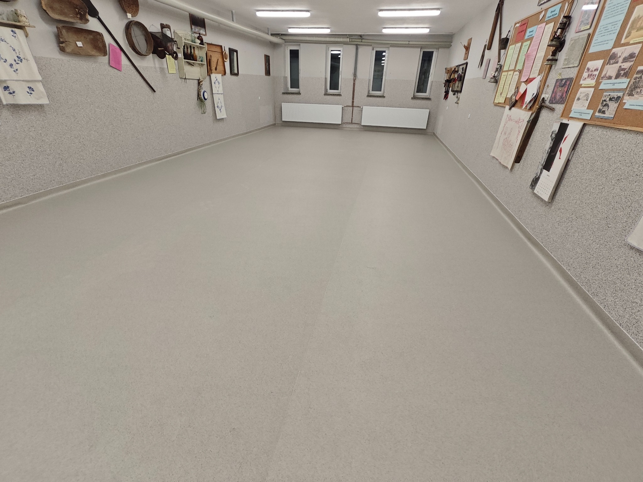 Jasnoszara, jednolita wykładzina podłogowa w przestronnym pomieszczeniu z oknami i dekoracjami na ścianach. Widoczne listwy przypodłogowe i grzejniki.