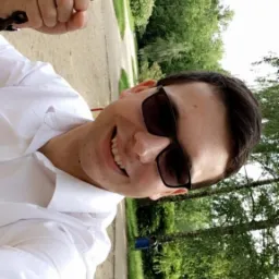 Uśmiechnięty mężczyzna w okularach przeciwsłonecznych, ubrany w białą koszulę, trzyma się łańcucha huśtawki na tle parku z drzewami i ścieżką.