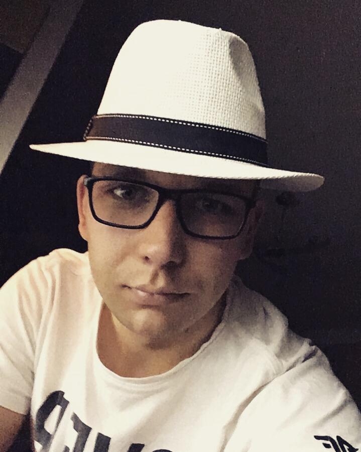 Portret mężczyzny w okularach i białym kapeluszu z czarną wstążką, ubrany w białą koszulkę z nadrukiem, na ciemnym tle.