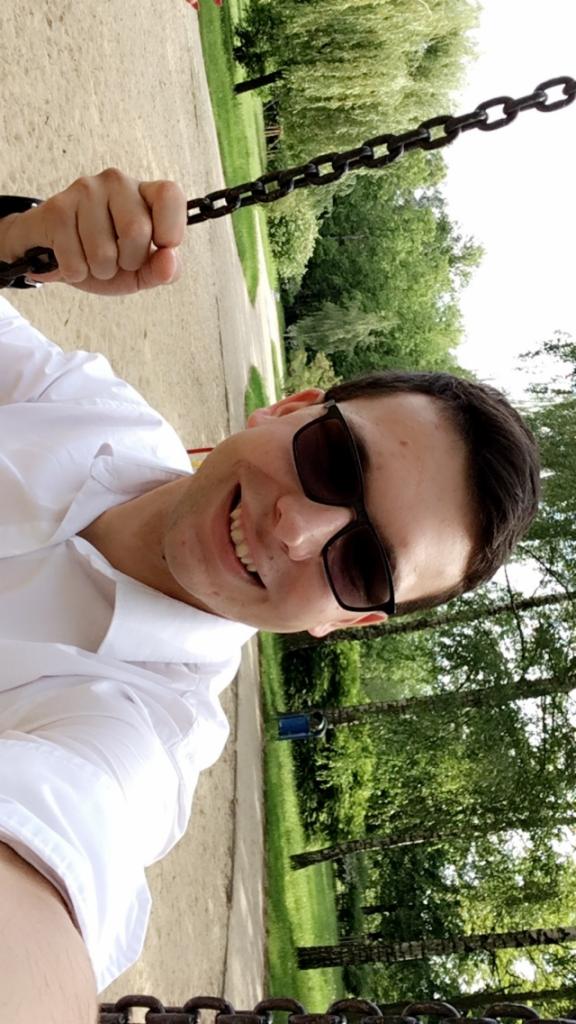 Uśmiechnięty mężczyzna w okularach przeciwsłonecznych, ubrany w białą koszulę, trzyma się łańcucha huśtawki na tle parku z drzewami i ścieżką.