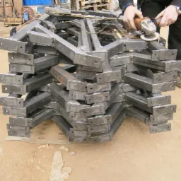 Stos surowych, metalowych elementów konstrukcyjnych w kształcie litery L, przygotowywanych do spawania, szlifowanych szlifierką kątową przez pracownika w roboczym ubraniu.