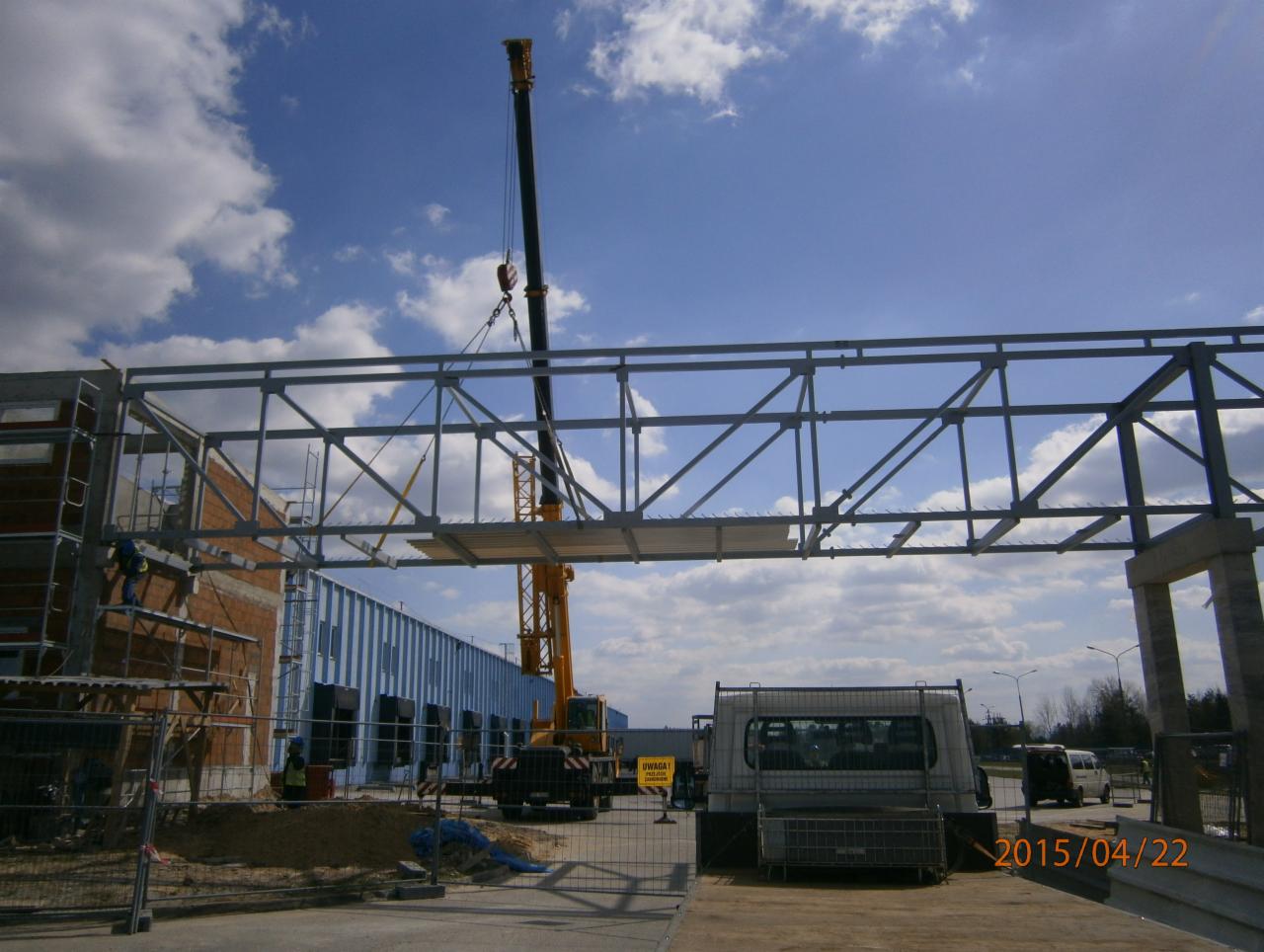 Montaż stalowej konstrukcji hali z użyciem dźwigu, widoczny fragment budynku w trakcie budowy i samochód dostawczy na placu budowy.