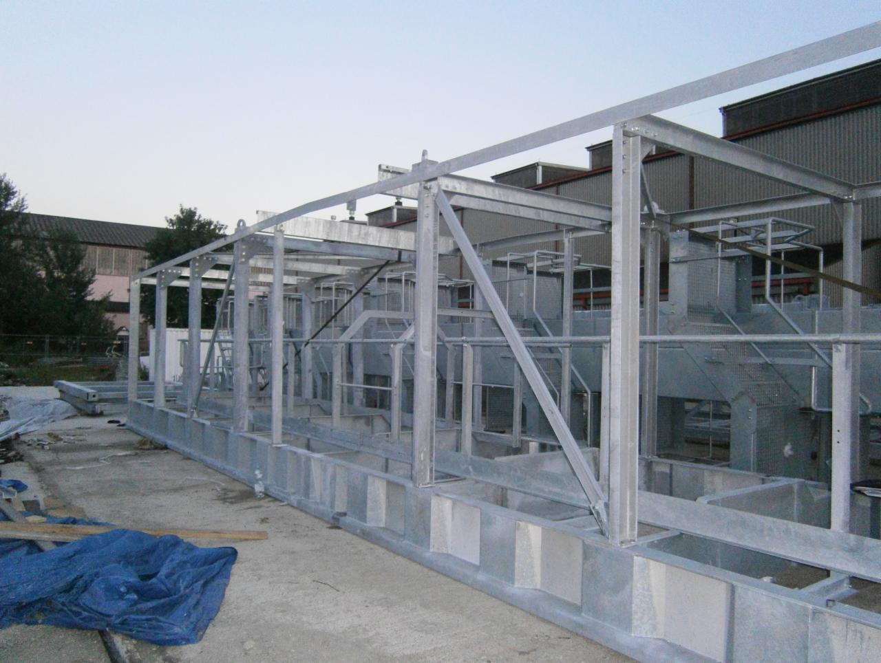 Szkielet stalowej konstrukcji hali przemysłowej w trakcie montażu, widoczne elementy nośne i łączenia skręcane, na zewnątrz budynku przemysłowego, z niebieską plandeką u dołu.