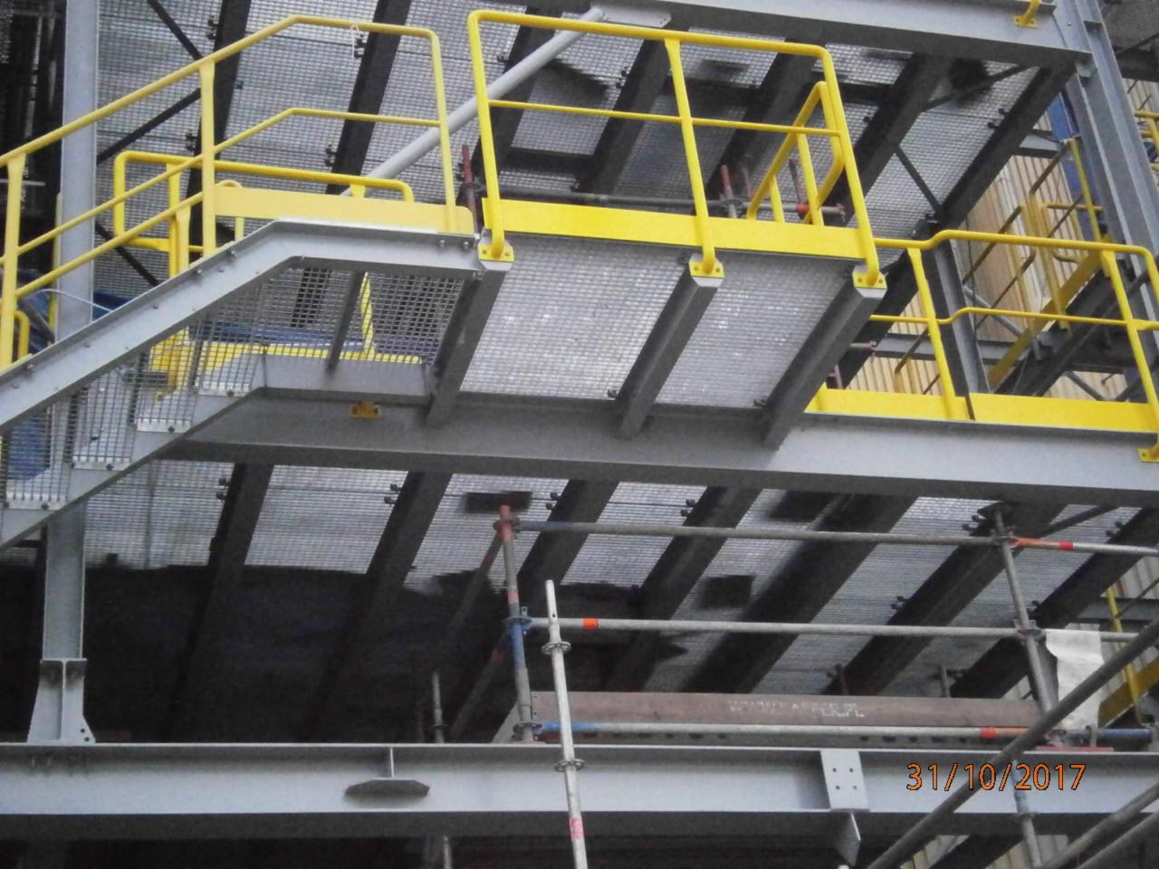 Stalowa konstrukcja platformy roboczej z żółtymi poręczami, widok z dołu na kraty pomostowe i elementy rusztowania w tle.