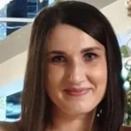 Uśmiechnięta brunetka w bluzce na tle jasnego wnętrza z elementami dekoracyjnymi. Portret kobiety o pogodnym wyrazie twarzy, z delikatnym makijażem i naturalnym oświetleniem.