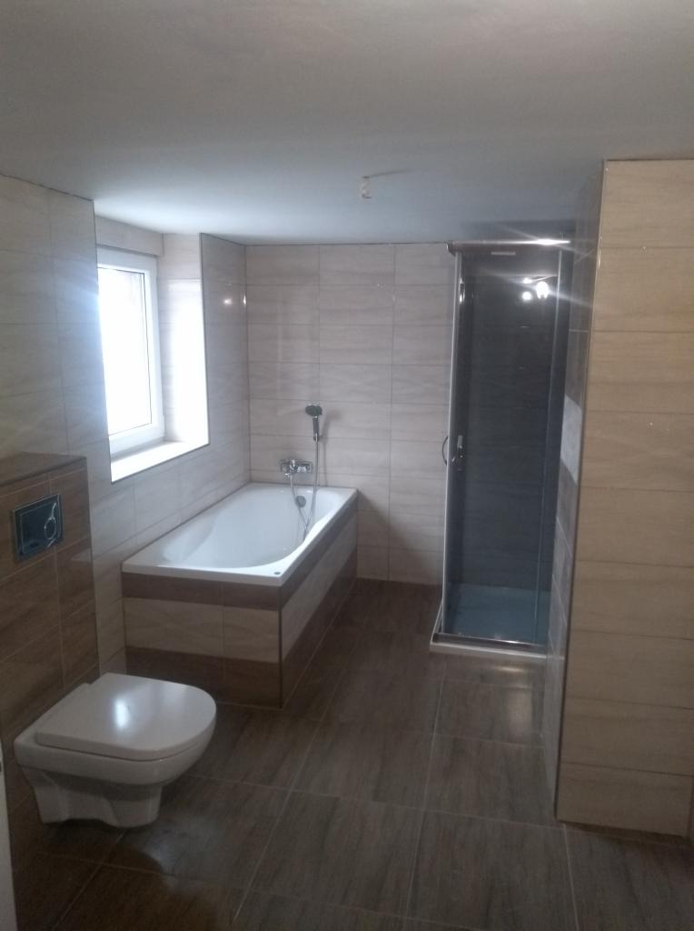 Wykończona łazienka z beżowymi płytkami imitującymi drewno, kabiną prysznicową z ciemnego szkła, białą wanną obudowaną płytkami oraz toaletą z zabudowaną spłuczką. Widoczne okno z białymi ramami.