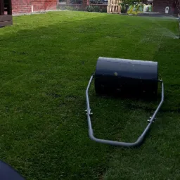 Świeżo założony trawnik z rolki, ubijany walcem ogrodowym. W tle widoczne zraszacze i paleta z workami ziemi.