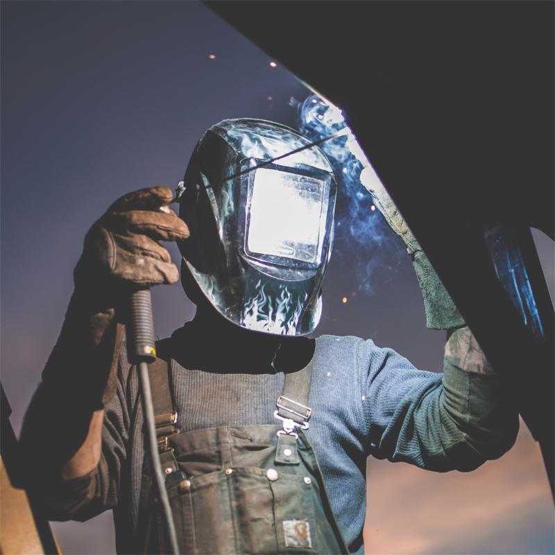 Spawacz w kombinezonie roboczym i masce ochronnej, pracujący nad konstrukcją stalową, widoczne iskry i dym.