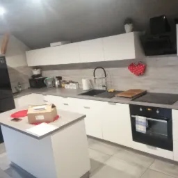 Nowoczesna kuchnia w odcieniach bieli i szarości z wyspą kuchenną, na której leży kartonowe pudełko, widok na płytki podłogowe i sprzęty AGD.