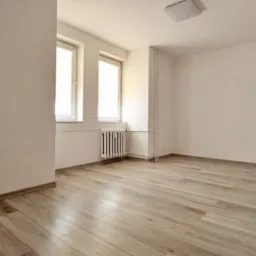 Jasne wnętrze pokoju z panelami podłogowymi o wzorze drewna, białymi ścianami, grzejnikiem i dwoma oknami w białych ramach, oświetlone przez lampę sufitową.