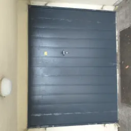 Szara, pozioma brama garażowa z centralnym uchwytem, zamontowana w jasnej elewacji budynku, obok widoczna lampa zewnętrzna i fragment chodnika z liśćmi.