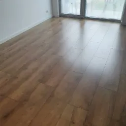 Ułożona podłoga z paneli imitujących drewno w jasnym pomieszczeniu z dużym oknem, widoczne listwy przypodłogowe i dekoracyjne elementy na ścianie.