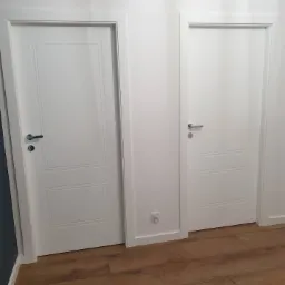 Dwa białe, lakierowane skrzydła drzwiowe z frezowanymi panelami, zamontowane w białych ościeżnicach na tle białej ściany i podłogi z paneli imitujących drewno, widoczne chromowane klamki i gniazdko...