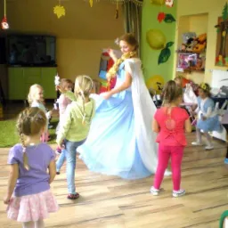 Animator w stroju księżniczki tańczy z grupą dzieci w udekorowanej sali. Zwraca uwagę jasna suknia i warkocz postaci.