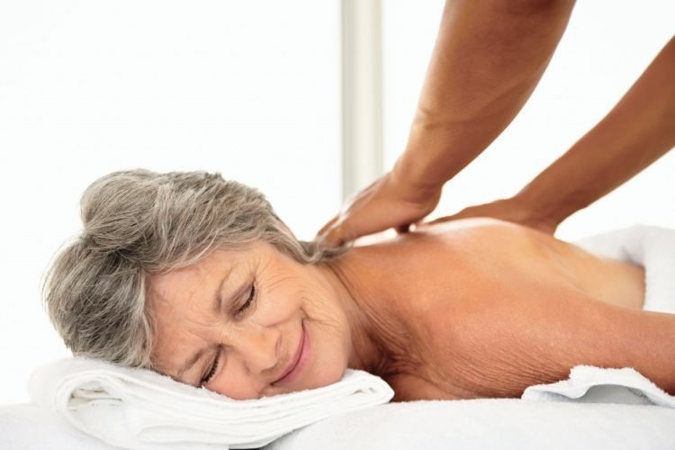 Starsza kobieta z szarymi włosami relaksuje się podczas masażu pleców, leżąc na białym ręczniku, z zamkniętymi oczami i delikatnym uśmiechem.
