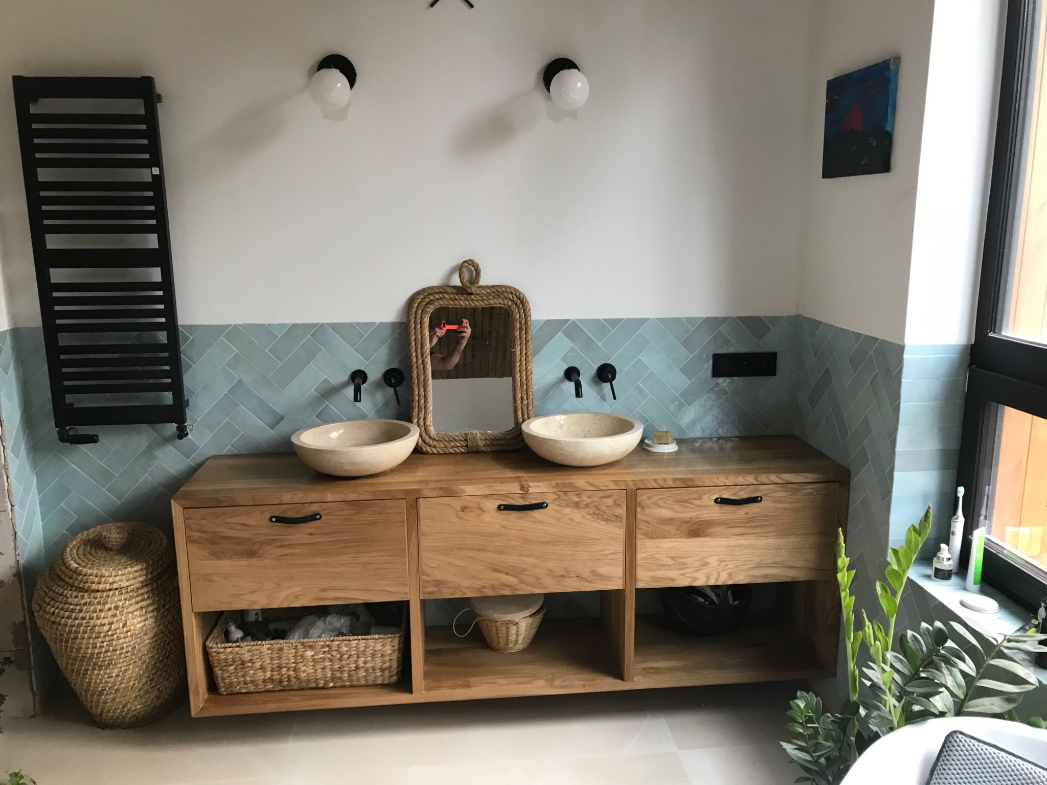 Łazienka z dwiema okrągłymi umywalkami nablatowymi z kamienia, drewnianą szafką z szufladami i otwartymi półkami, lustrem w ramie ze sznura oraz niebiesko-szarą glazurą ułożoną w jodełkę na ścianie.