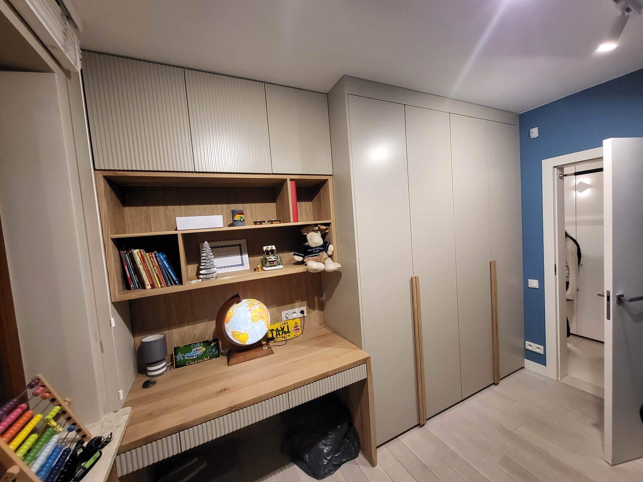 Pokój dziecięcy z biurkiem z drewnianym blatem, regałem i szafą w zabudowie w odcieniach szarości, z globusem i zabawkami na półkach, oraz abakusem na biurku.