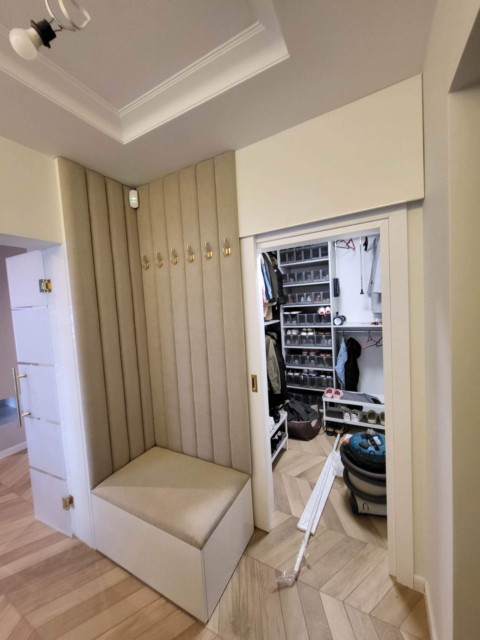 Przedsionek z tapicerowanym siedziskiem i panelem ściennym z wieszakami, widok na garderobę z półkami na buty i ubrania.