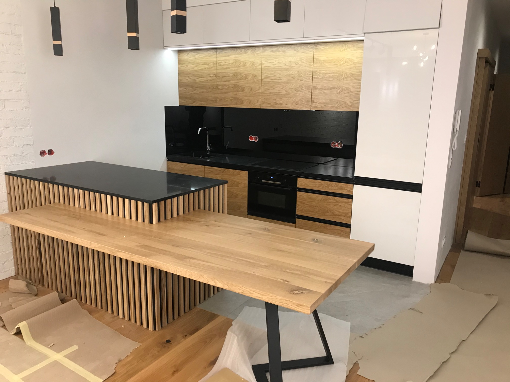 Nowoczesna kuchnia z drewnianymi frontami szafek i czarnym blatem, wyspa kuchenna z drewnianymi lamelami i drewnianym stołem na metalowej podstawie, widoczne zabezpieczenia podłogi na czas montażu.