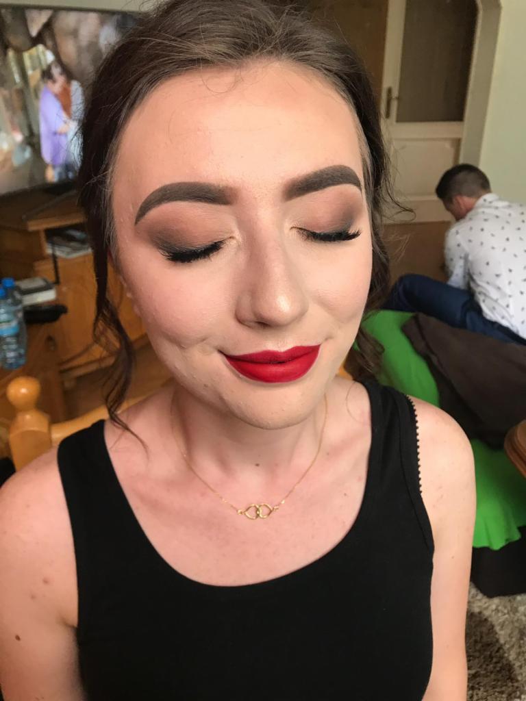 Portret kobiety z zamkniętymi oczami, prezentującej wykonany dzienny make-up z matowymi cieniami w odcieniach brązu, podkreślonymi brwiami i intensywnie czerwoną pomadką na ustach. Na szyi widoczny...