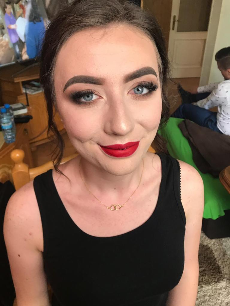 Portret kobiety z intensywnym makijażem oczu i ust, uśmiechająca się, w tle widoczny mężczyzna