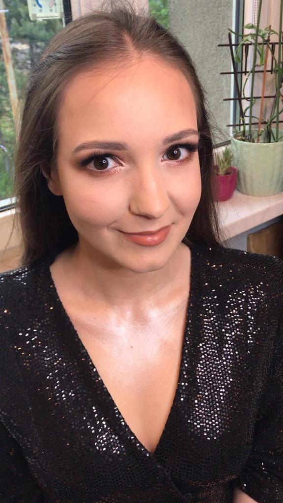 Portret uśmiechniętej brunetki z delikatnym makijażem oczu i ust w odcieniach brzoskwini, ubrana w czarną sukienkę z cekinami, na tle okna z roślinami doniczkowymi.