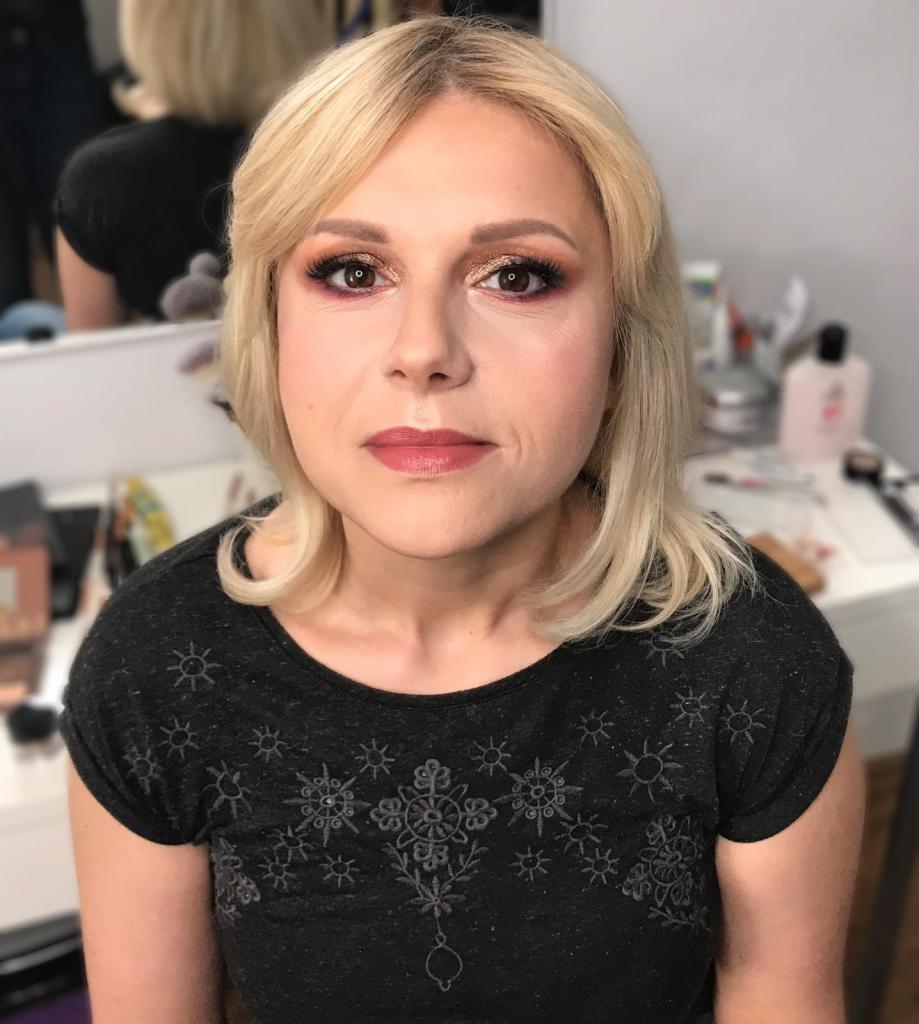 Portret kobiety z blond włosami i delikatnym, rozświetlającym wizażem, z uwzględnieniem złotych cieni na powiekach i subtelnie podkreślonych ustach. W tle widoczne kosmetyki na toaletce.