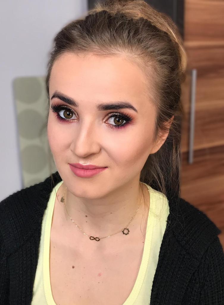 Portret kobiety z delikatnym, różowym błyszczykiem na ustach i starannie wykonanym cieniowaniem powiek w odcieniach złota i fioletu, uzupełnionym długimi, gęstymi rzęsami. Zauważalny złoty...