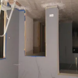 Wnętrze w trakcie przygotowań do odświeżenia ścian, zabezpieczone taśmą malarską wokół okien i otworów, widoczna drabina aluminiowa i wstępnie pomalowane ściany na szaro.