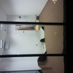 Widok przez okno na wnętrze biura w trakcie prac wykończeniowych, z częściowo położoną wykładziną dywanową, zabezpieczoną folią i widoczną drabiną w tle.