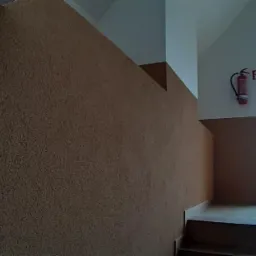 Wnętrze klatki schodowej z brązową, ziarnistą ścianą, widokiem na zewnątrz przez okno i gaśnicą na ścianie.