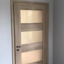 Zamknięte drzwi wewnętrzne z czterema poziomymi szybami z matowego szkła, o ramie w kolorze jasnego drewna, zamontowane w szarej ścianie z widoczną listwą przypodłogową.