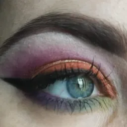 Zbliżenie na oko z intensywnym makijażem: fioletowo-różowe cienie na powiece, pomarańczowa kreska, czarny eyeliner w stylu cat-eye, zielone akcenty na dolnych rzęsach, widoczne niedoskonałości skóry.