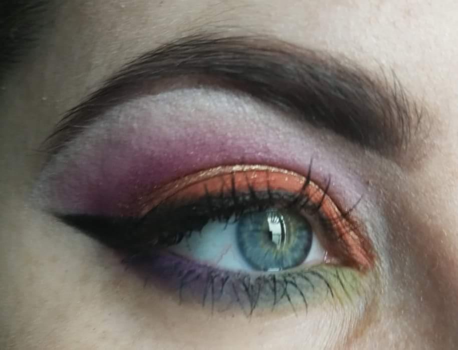 Zbliżenie na oko z intensywnym makijażem: fioletowo-różowe cienie na powiece, pomarańczowa kreska, czarny eyeliner w stylu cat-eye, zielone akcenty na dolnych rzęsach, widoczne niedoskonałości skóry.