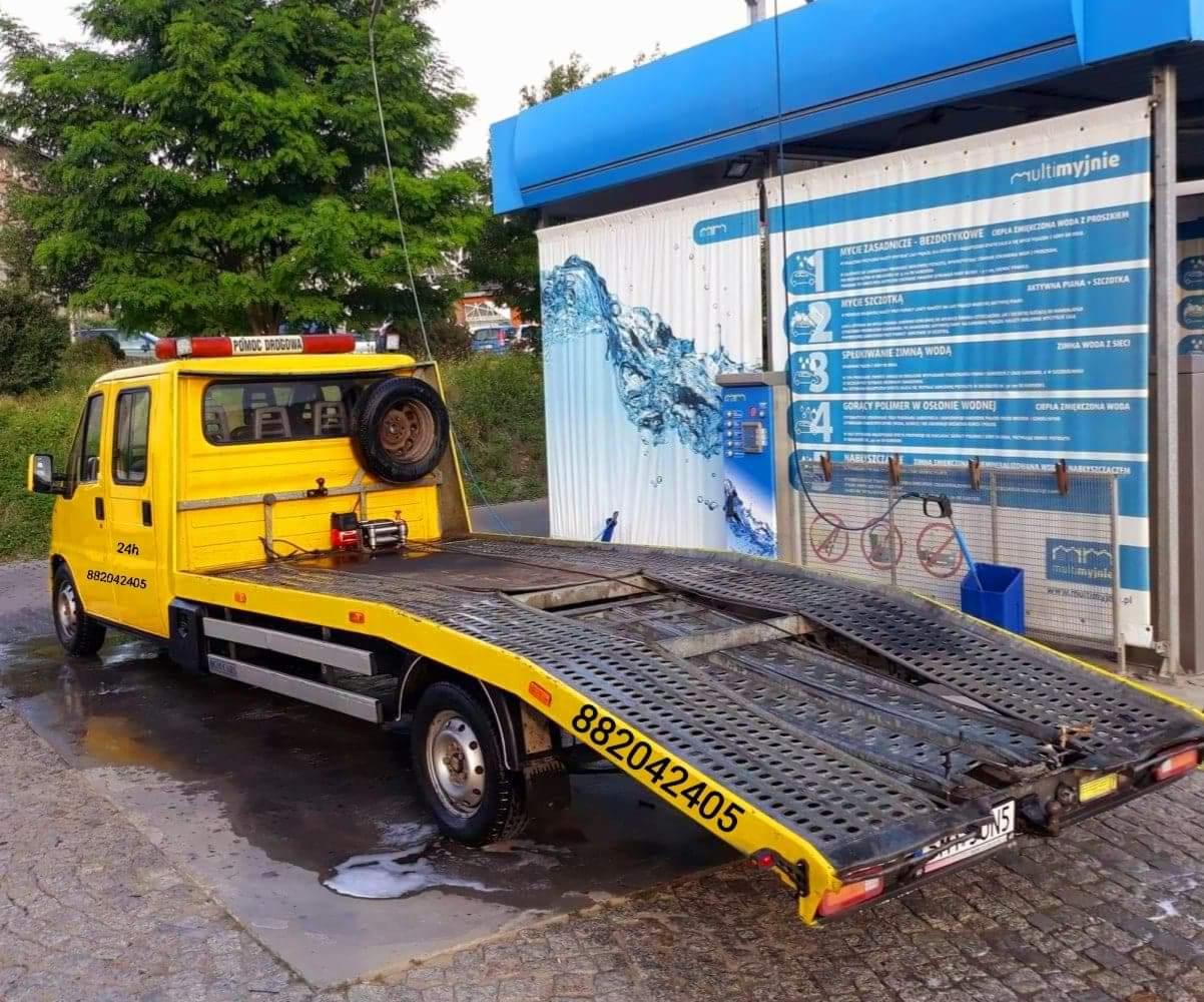 Żółty pojazd pomocy drogowej z platformą załadunkową stoi przed myjnią samochodową, widoczny napis 'Pomoc Drogowa' i numer telefonu