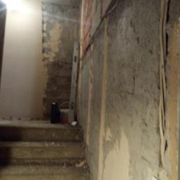 Olbud - Klatka schodowa w trakcie remontu: surowe ściany z pozostałościami tynku, widoczne przewody elektryczne, poziomica oparta o ścianę, wiadro i pojemnik na schodach, przygotowanie do dalszych prac...