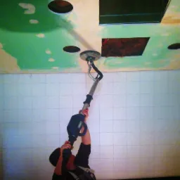 Szlifierka do gipsu na wysięgniku w trakcie pracy przy suficie z otworami na oświetlenie i częściowo pomalowanym na zielono, ściana wyłożona białymi płytkami.