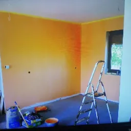 Pokój w trakcie malowania na pomarańczowo-żółty odcień, widoczna drabina, wiadro z farbą i worek z gładzią Knauf MP 75, zabezpieczona taśmą krawędź sufitu, przygotowane otwory pod gniazdka.